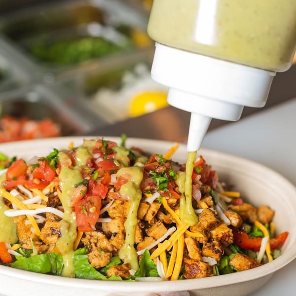 A squeeze bottle drizzles green dressing over a Mexican chicken salad bowl with lettuce, cheese, and pico de gallo.