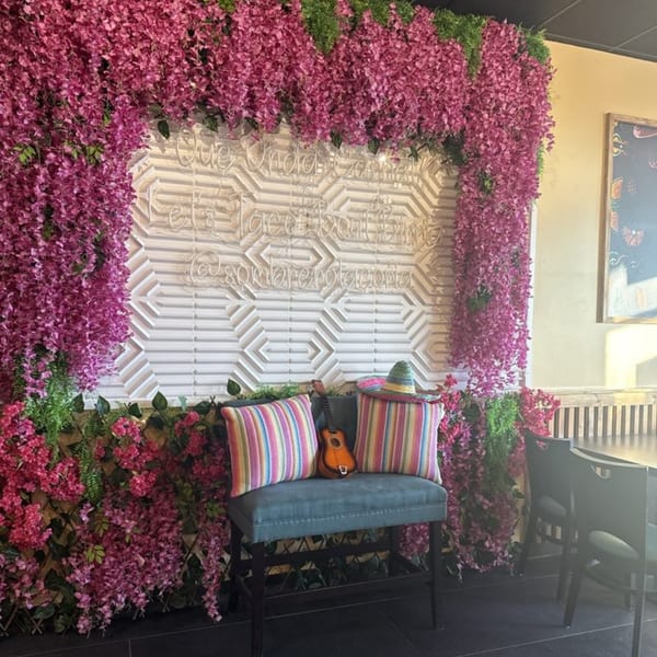 A vibrant floral wall with pink and green flowers, a neon sign, and a blue bench with colorful pillows.