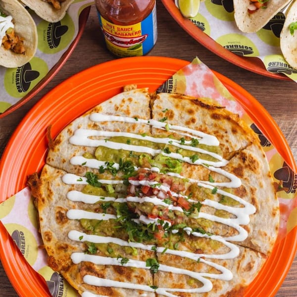 A large quesadilla with guacamole, sour cream, and pico de gallo, alongside tacos and hot sauce.