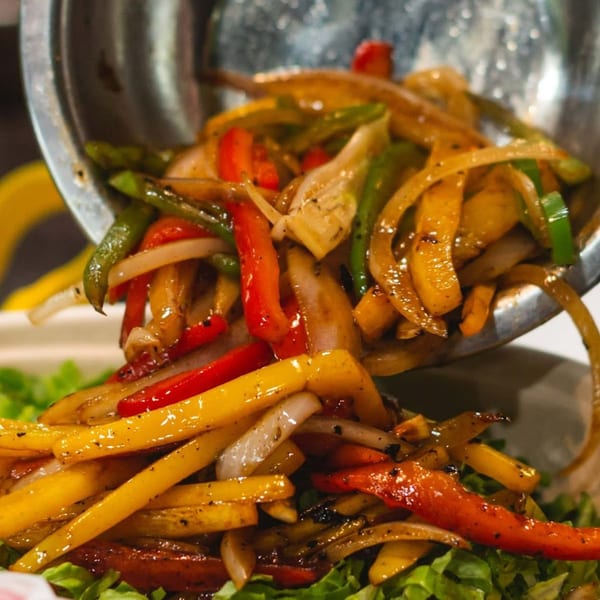 Sizzling red, green, and yellow bell peppers and onions being poured from a metal bowl.