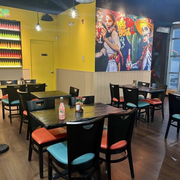Restaurant interior with yellow walls, colorful mural, dark tables, and chairs with orange and blue cushions.
