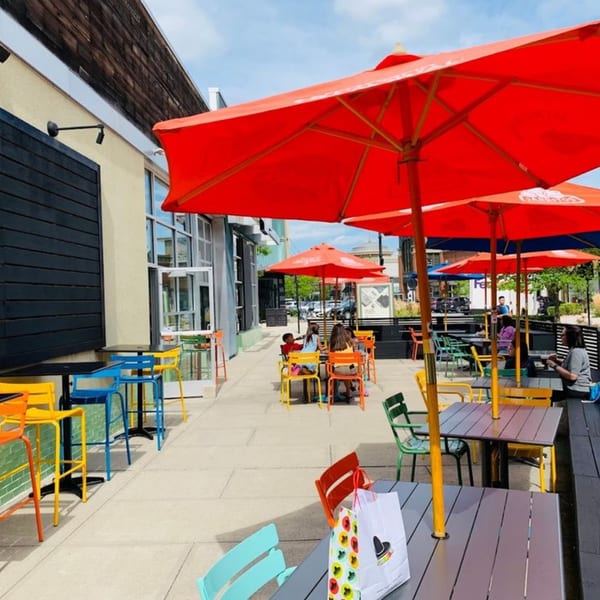 Outdoor patio with red umbrellas, colorful chairs, and people dining at tables on a sunny day.