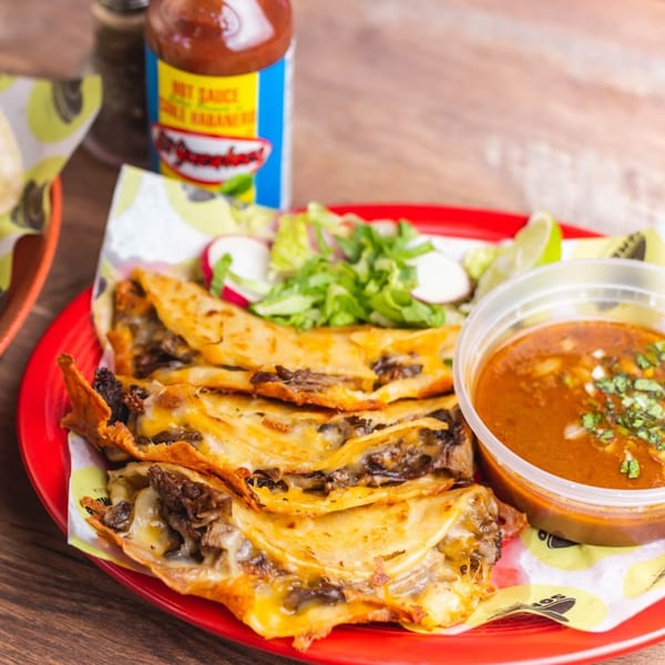 Three cheesy birria tacos with a side of consommé, salad, and lime on a red plate.