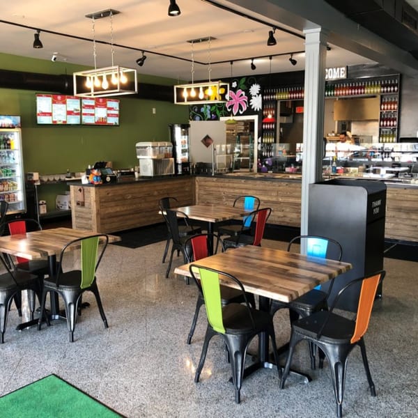 Modern restaurant interior with wooden tables, colorful chairs, and a counter with menu screens.