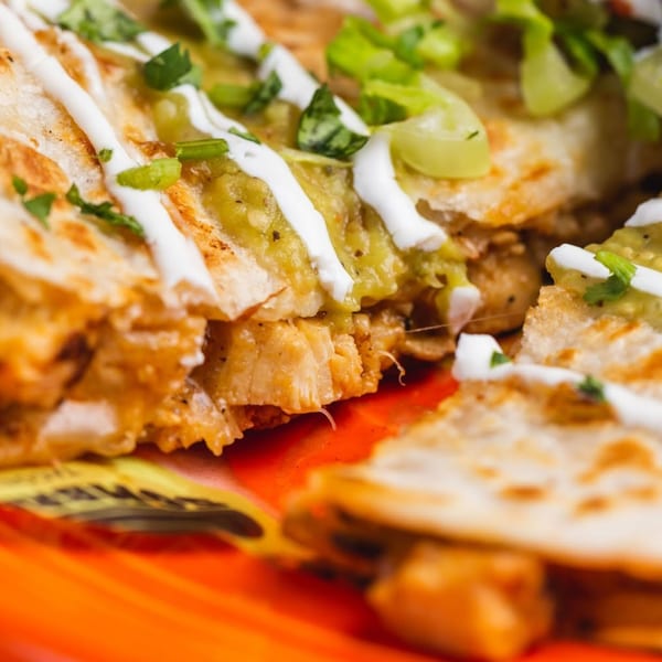 Close-up of chicken quesadillas topped with guacamole, sour cream, and green onions on an orange plate.