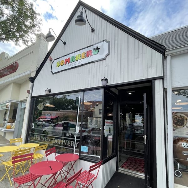 The exterior of Sombrero Tacoria, a white building with a black roof, and red and yellow outdoor seating.
